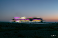 La Croisière en couleurs — navire aux lumières roses, jaunes et blanches étirées au crépuscule, silhouettes sur le rivage | Bybadr