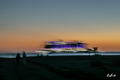 La Traversée violette — navire aux lumières violettes et blanches sous un ciel orangé, silhouettes sombres sur le rivage | Bybadr